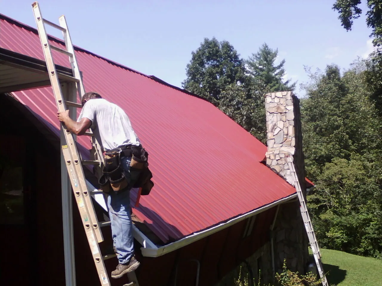 Roofer on a ladder beside a red metal roof and stone chimney — Soffit & Fascia Repair by Roofing Brothers in Winooski